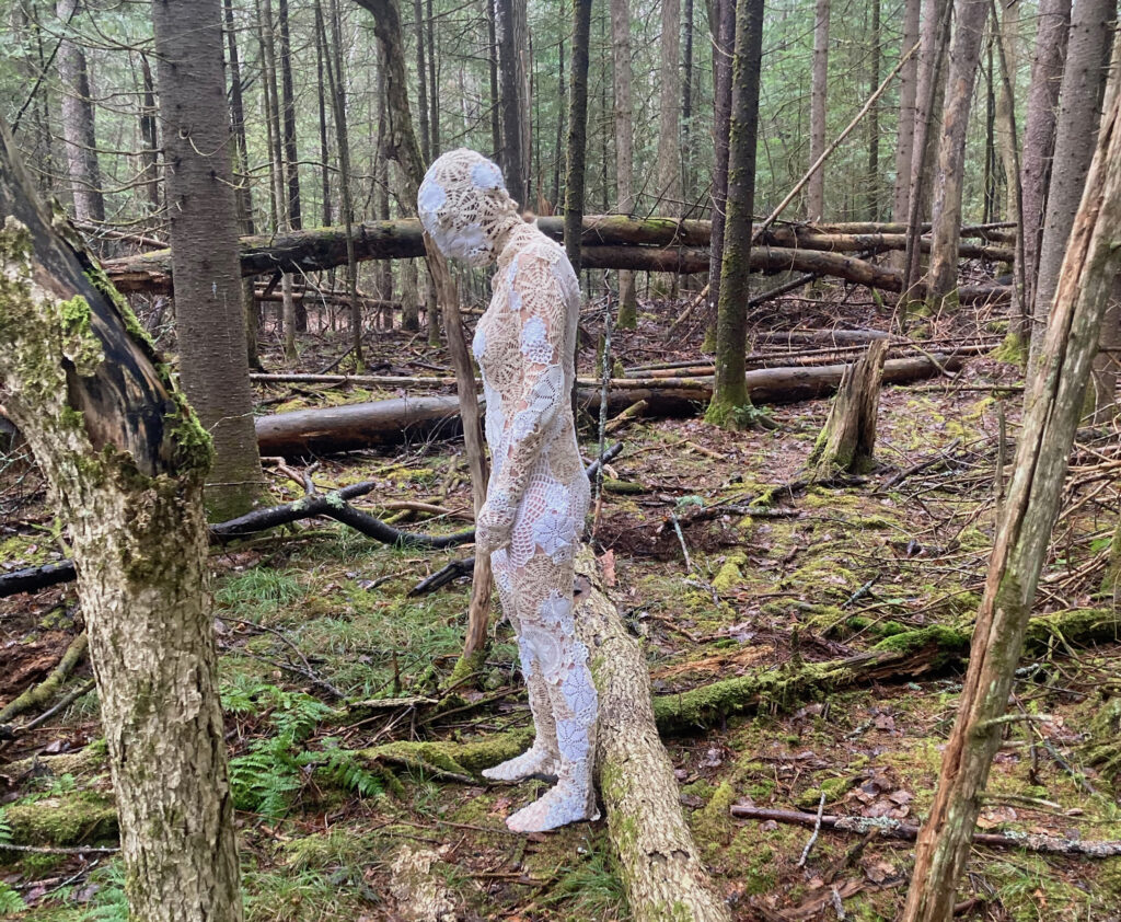 Form of a woman in a bodysuit fashioned of various pieces of found lace, and lace doilies standing alone in a spruce forest