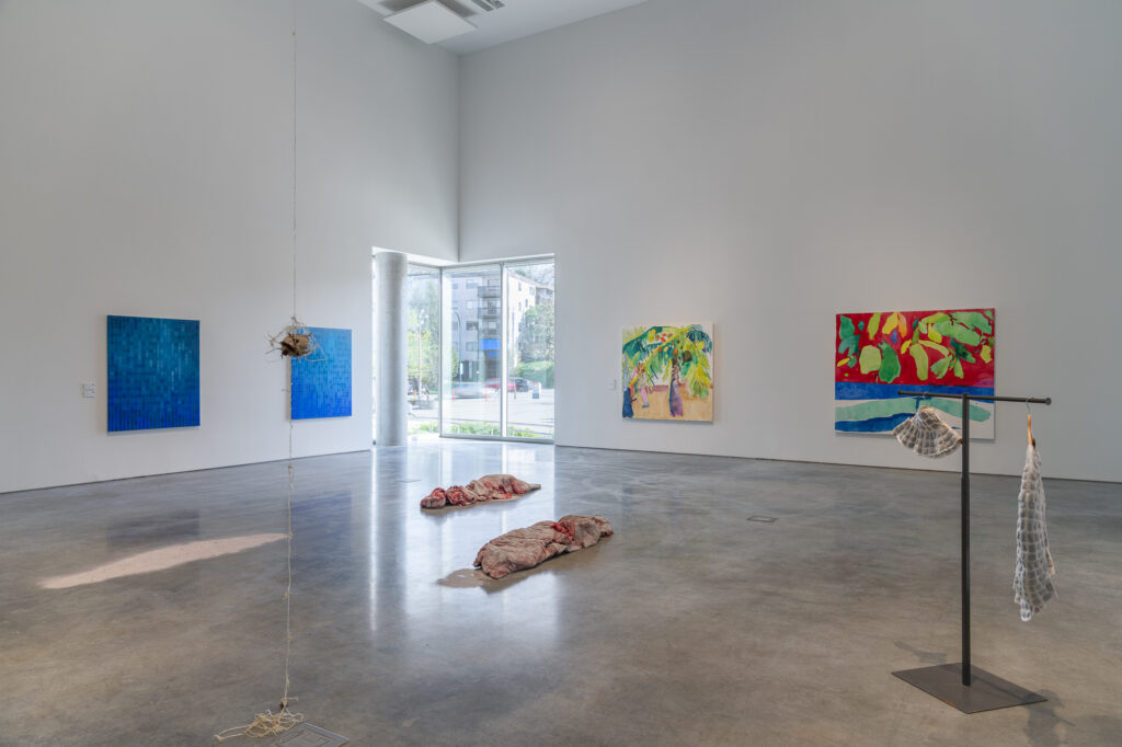 An exhibition space located near the window, showing a round object hanging in the middle of the room from a string that descends from the ceiling. To the right of it is a clothing stand with a grey block-like patterned hat and shirt. In the left background are two blue gradient paintings created by tiny square blocks. To the right are two paintings of tree-like illustration in vibrant shades.
