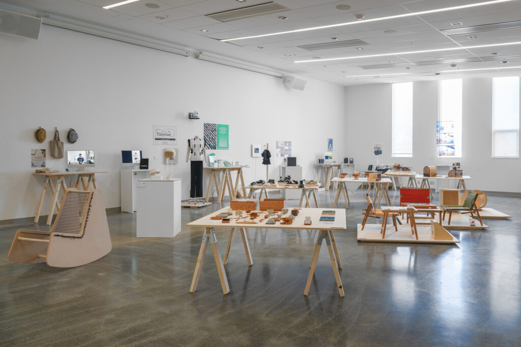
A view of the interior space in Emily Carr University, where the central area features a collection of chair designs, along with crafted shoes and smaller ceramic objects on display tables. On the left side, various projects are showcased, including hats and costumes on mannequins, with posters on the wall and computers screening videos and images.