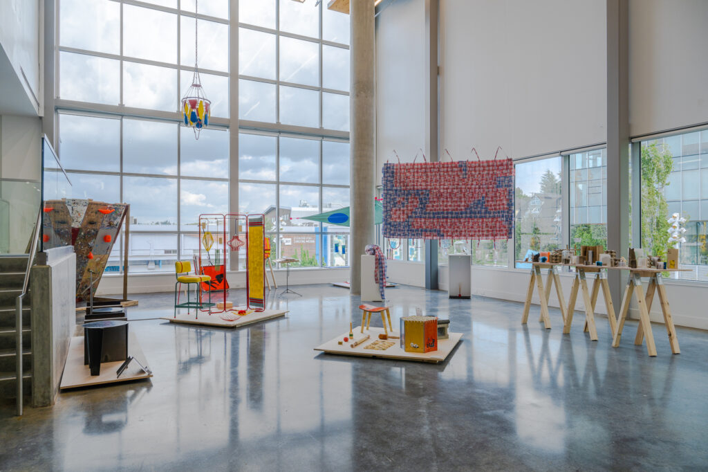 Inside an interior window view of Emily Carr University, two collections of furniture designs in bright shades of red and yellow sit in the center, with a rectangular graphic featuring a red and blue woven pattern hanging prominently in the background. On the right, a series of smaller display objects are placed on a wooden table stand.