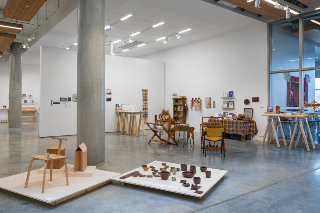 In an exhibition space, a series of wooden furniture designs sit in the front, with a collection of brown ceramics spread out on the floor installation stand to the right. In the background, two wooden loom weaving machines are visible, while the far background features more handcrafted projects placed on the wall and on table stands.