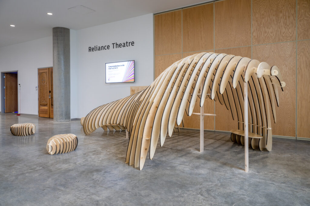 A wooden installation with a long, curvy architectural shape that includes bench seating inside and three individual seats positioned to the left of the installation.