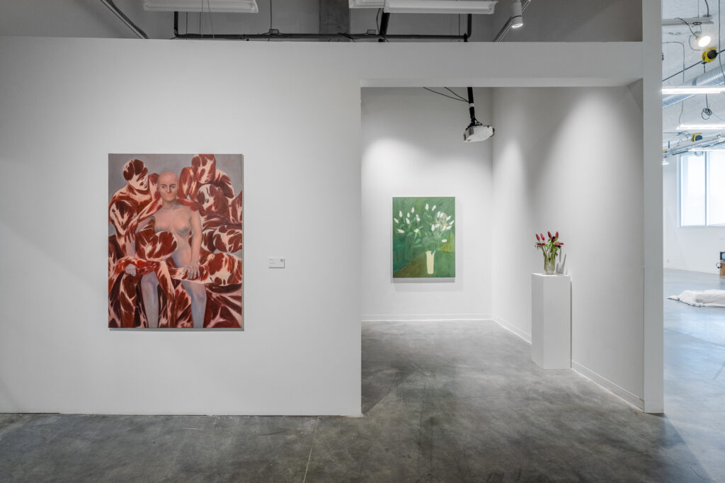 Exhibition installation inside the Emily Carr university building. In the foreground is an oil painting of a nude human surrounded by flesh-painted human figures. On the far wall inside a cut-open room is a green painting of white flowers in a cylinder vest. To the right of the painting is a physical display of a bouquet of red flowers.