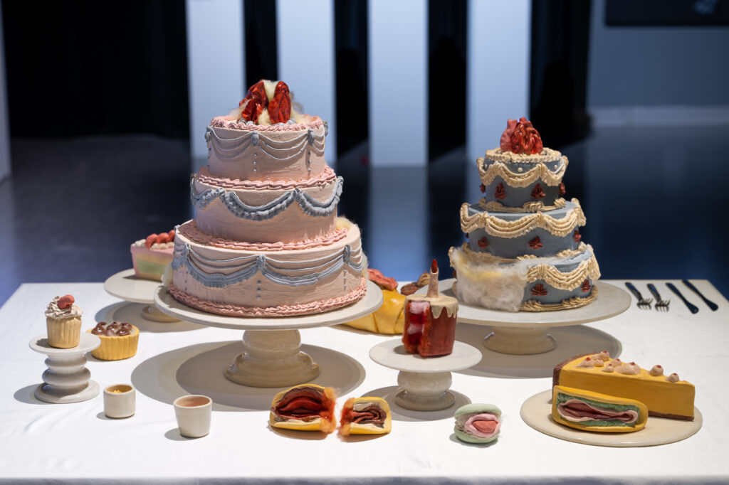 On a flat, white installation table, there is a scene of desserts and cakes made from ceramic and wool, featuring two three-layered large cakes in the center, surrounded by small desserts, cupcakes, and utensils.