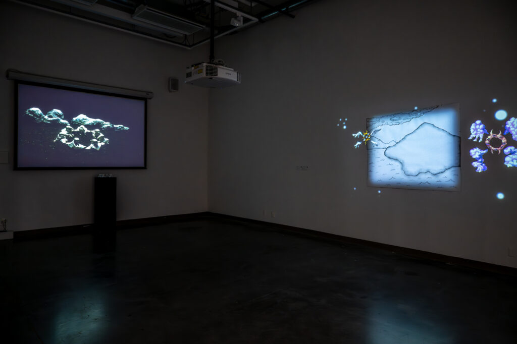 In a dimly lit room, a projector fixed to the ceiling is projecting two artworks - on the left is a scene of seven objects with a physical model on a stand below the projector screen, and on the right is a black and white sketch with an abstract illustration overlaid on top.