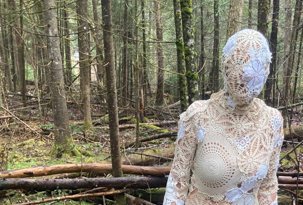 Form of a woman in a bodysuit fashioned of various pieces of found lace, and lace doilies standing alone in a spruce forest
