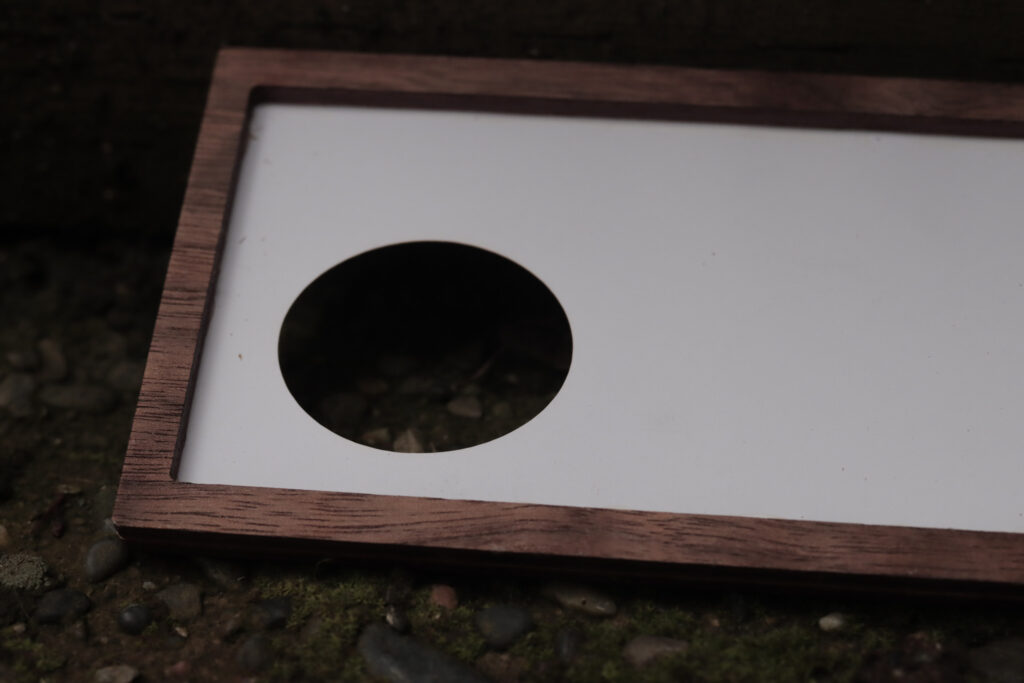 Wooden frame with a paper insert that has a small circle cut out on the lower left corner, laying on a stone surface 