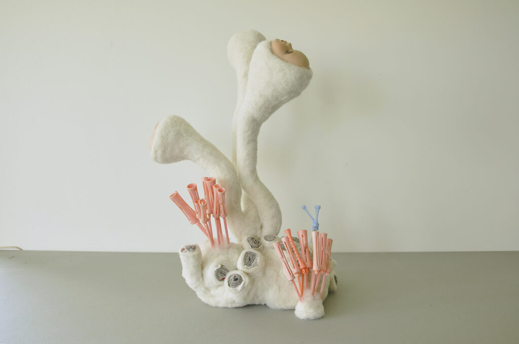 A sculpture resembling coral, consisting of three white needle felted forms with porcelain doll faces, pink and blue disposable hair curlers and grey wool socks comprising smaller sections of coral 