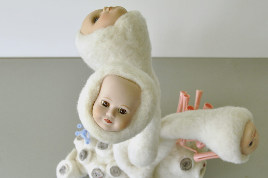 A detail view of a sculpture resembling coral, consisting of three white needle felted forms with porcelain doll faces, pink and blue disposable hair curlers and grey wool socks comprising smaller sections of coral 